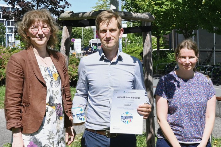 Zwei Männer und eine Frau stehen auf einer Wiese und lächeln und die Kamera. der Mann in der Mitte hält eine Urkunde in der Hand. 
