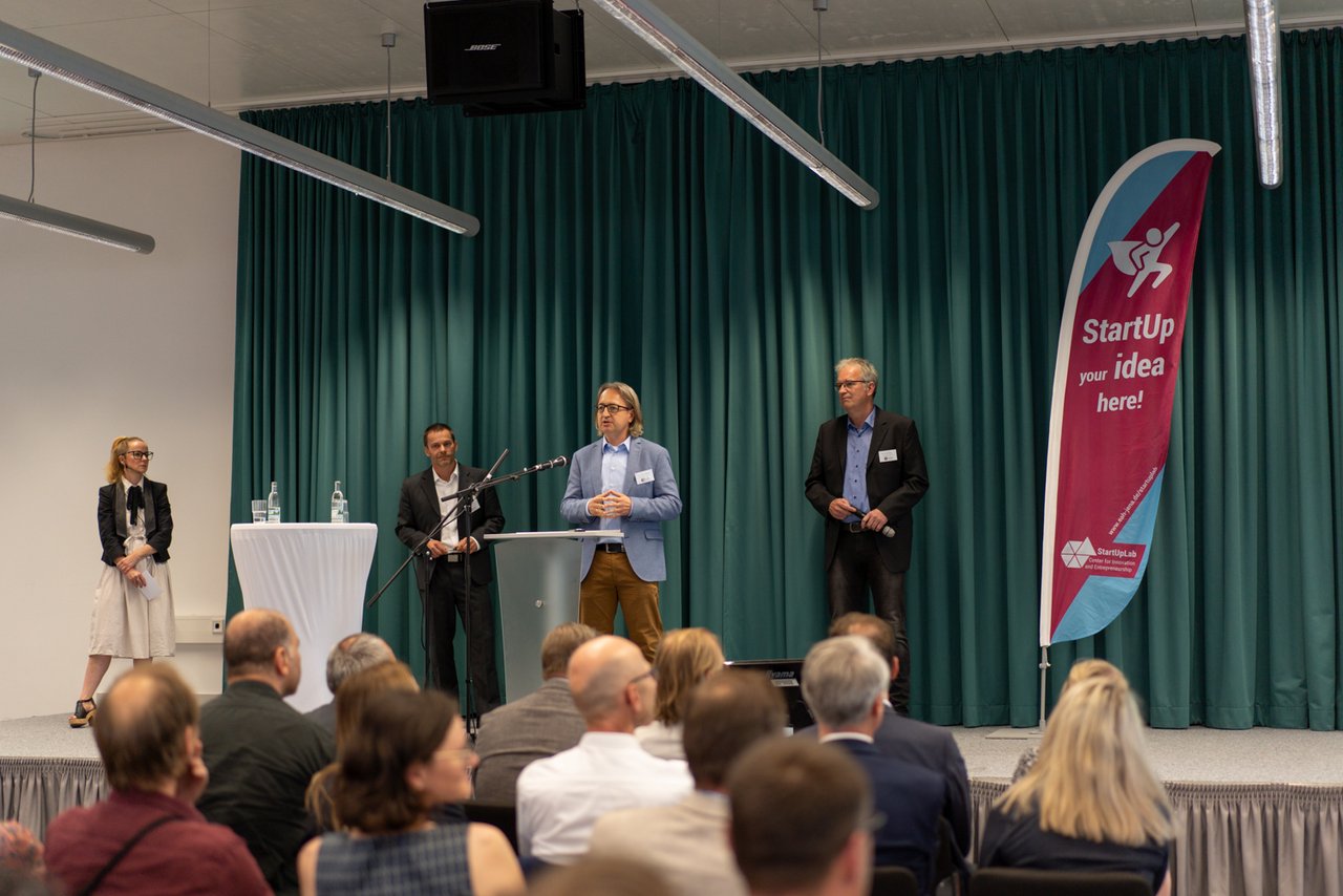 auf der Bühne stehen die drei Projektleiter und begrüßen die Veranstaltungsteilnehmenden
