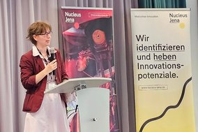 Frau am Stehpult mit Mikro in der Hand, im Hintergrund 2 Rollups des Projekts "Nucleus Jena".