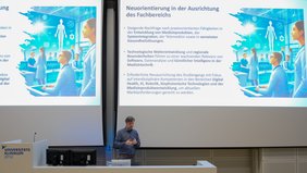 Eine Person, in Halbtotale, referiert vor einer Leinwand in einem Hörsaal.