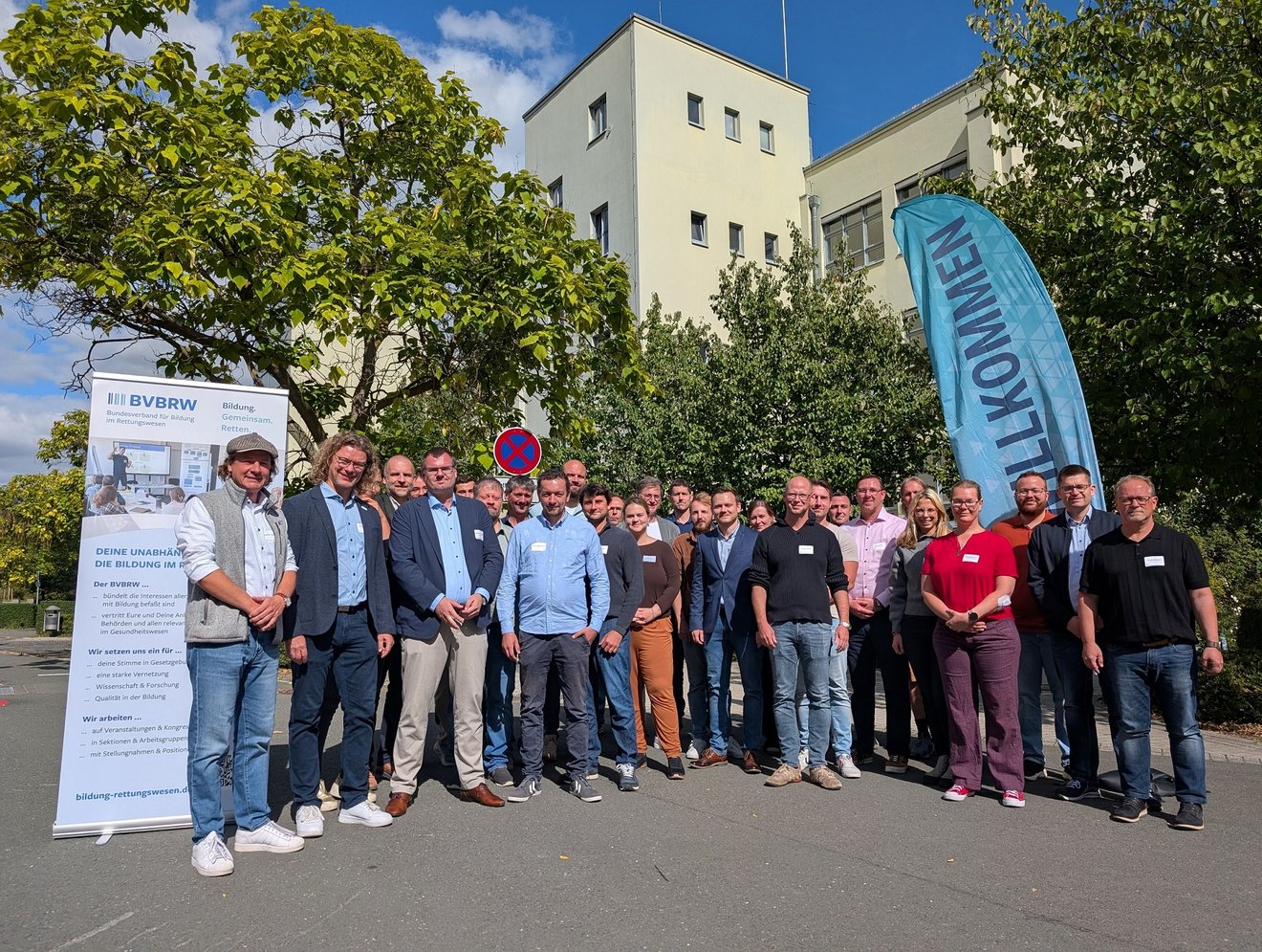 Eine Gruppe von ca. 30 Personen steht im Freien. Alle blicken zur Kamera. Im Hintergrund Bäume und ein großes Gebäude.