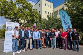 Eine Gruppe von ca. 30 Personen steht im Freien. Alle blicken zur Kamera. Im Hintergrund Bäume und ein großes Gebäude.