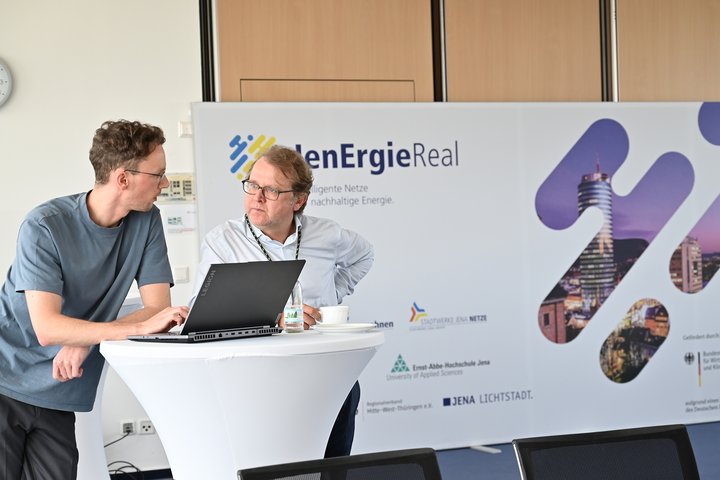 Zwei Männer stehen an einem Stehtisch und unterhalten sich, im Hintergrund ein großes Schild mit der Aufschrift JenEnergieReal