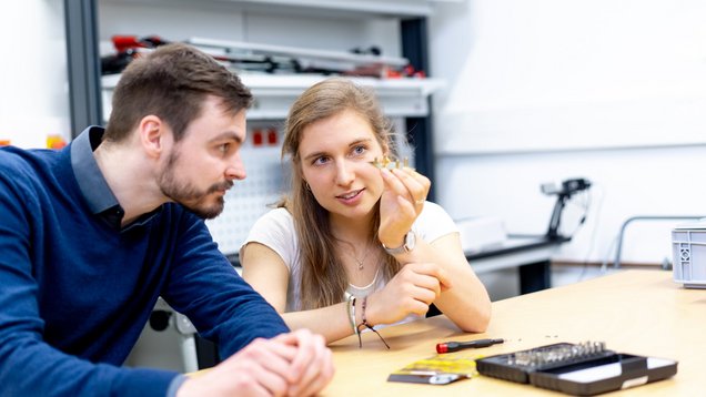 ein junger Mann und eine junge Frau sitzen an einem Tisch und schauen sich ein kleines Bauteil an