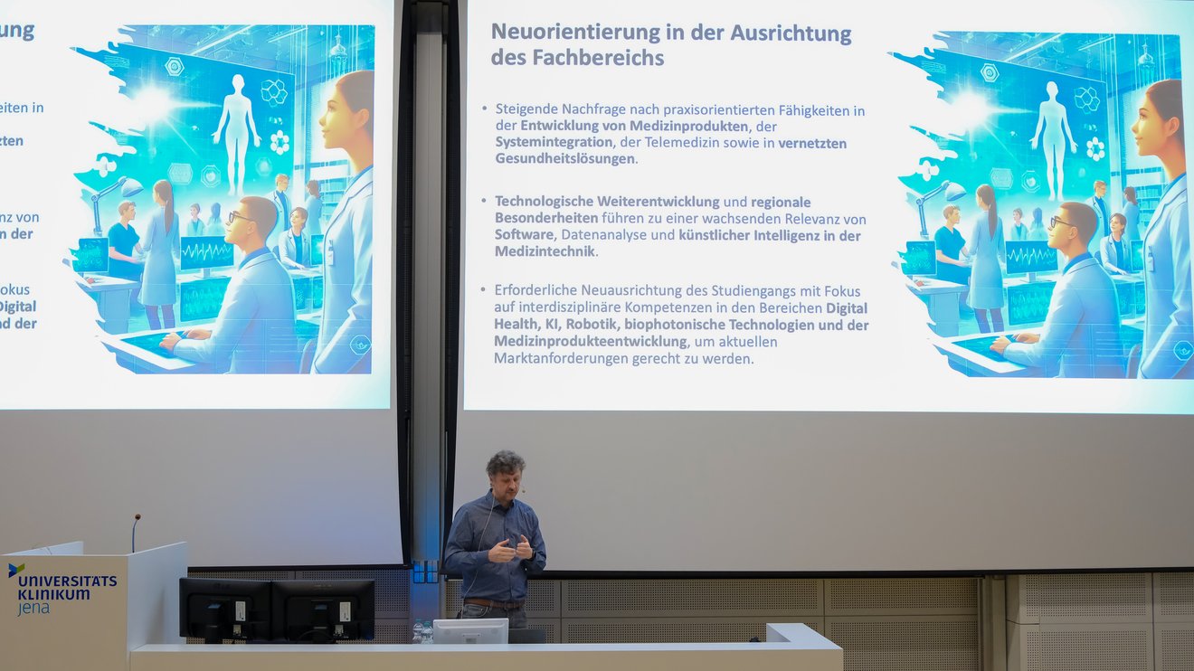 Eine Person, in Halbtotale, referiert vor einer Leinwand in einem Hörsaal.