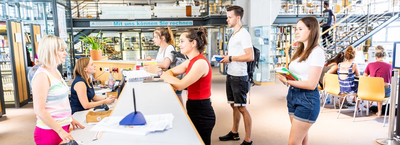 Studierende stehen am Tresen der Bibliothek um Bücher auszuleihen.