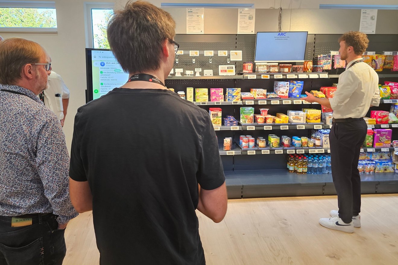 Mehrere Personen stehen in einem Raum vor einem Regal mit Lebensmitteln und weiteren Produkten. Eine Person erklärt die Technik im Raum.