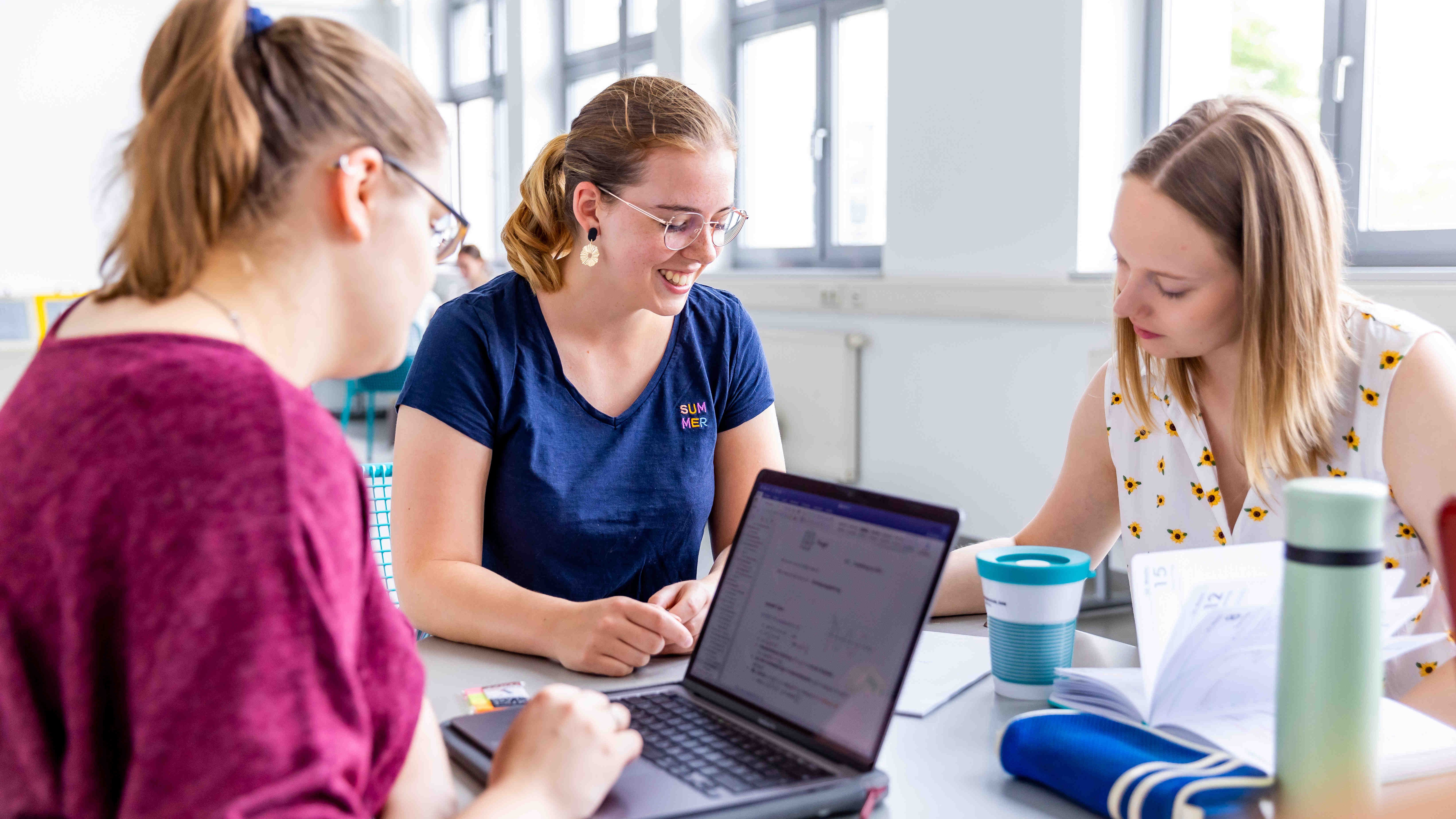 Drei junge Frauen sitzen an einem Tisch, eine davon arbeitet am Laptop.