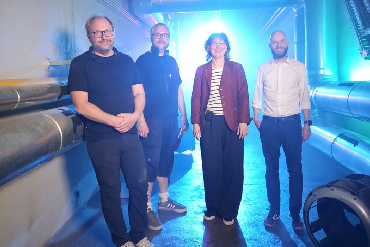 Drei Männer und eine Frau stehen in einem Kellergang und blicken in die Kamera, rechts und links sind Rohre an den Wänden, von hinten strahlt ein blaues Licht.