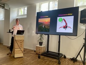 Ein Referent steht hinter einem Pult links neben einem Smartboard und spricht.