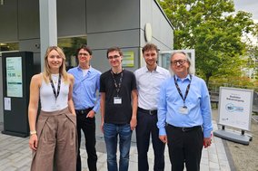 Eine Frau und 4 Männer stehen vor einem Gebäude und blicken in die Kamera