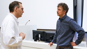 Zwei Personen unterhalten sich. Halbnahaufnahme im Profil. Im Hintergrund sieht man Monitore und ein Pult mit Mikro.