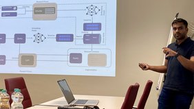 Eine Person steht in einem Seminarraum neben einer, an die Wand projizierten Powerpoint-Präsentation und spricht. 