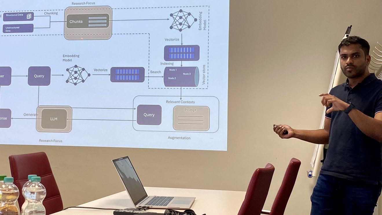Eine Person steht in einem Seminarraum neben einer, an die Wand projizierten Powerpoint-Präsentation und spricht. 