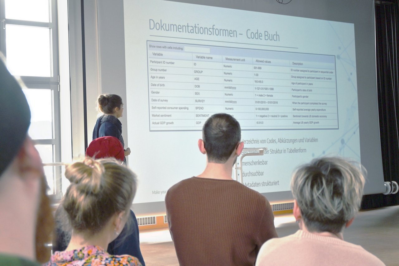 Impulsvortrag auf der FDM-Veranstaltung "Treasure your data" Ein Blick über die Schultern der Zuhörenden auf die Bühne mit der Referentin Dr. Cora Assmann beim Impulsvortrag zu guter Datendokumentation.