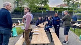Menschen sitzen und stehen im Freien an Bierbänken und unterhalten sich. Im Hintergrund wird gegrillt. 