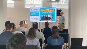 9 sitzende Personen in Rückansicht verfolgen einen Vortrag. Im Hintergrund eine referierende Person neben einer Präsentation.