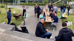 Eine Gruppe von zwanzig Personen beim Grillen aus Vogelperspektive. Menschen sitzen oder stehen an Bierbänken und unterhalten sich. 