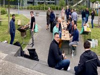 Eine Gruppe von zwanzig Personen beim Grillen aus Vogelperspektive. Menschen sitzen oder stehen an Bierbänken und unterhalten sich. 
