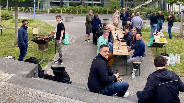 Eine Gruppe von zwanzig Personen beim Grillen aus Vogelperspektive. Menschen sitzen oder stehen an Bierbänken und unterhalten sich. 