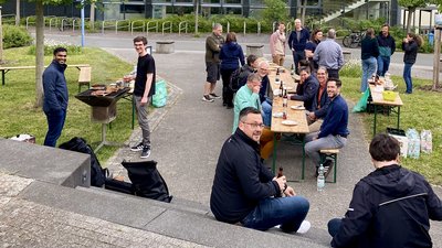 Eine Gruppe von zwanzig Personen beim Grillen aus Vogelperspektive. Menschen sitzen oder stehen an Bierbänken und unterhalten sich. 