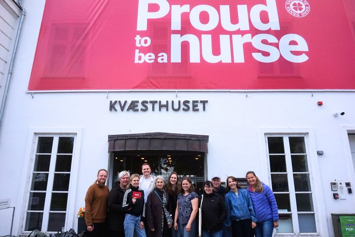 Gruppe von jungen Menschen, die vor einem Haus stehen. Am Haus hängt ein großes Banner, auf dem steht: Proud to be a nurse.