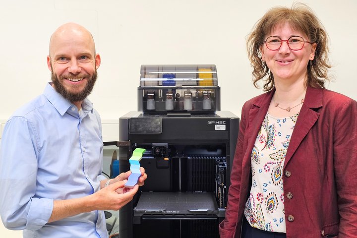 Ein Mann und eine Frau stehen vor einem 3D-Drucker und blicken in die Kamera. Der Mann hält ein kleines buntes 3D-getrucktes Teil in den Händen.