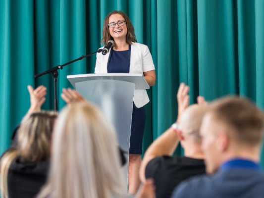 Eine Frau steht an einem Rednerpult. Vor ihr sitzen mehrere Menschen im Publikum.