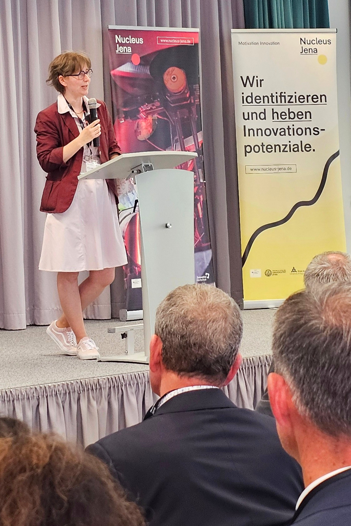 Frau am Stehpult mit Mikro in der Hand, im Hintergrund 2 Rollups des Projekts "Nucleus Jena".